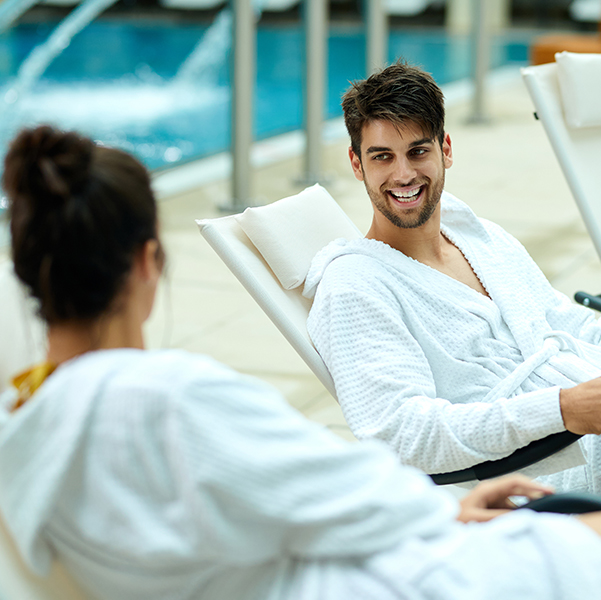 Young couple in bathrobes talking while sitting by the pool and spending a weekend at the spa. Focus is on happy man.